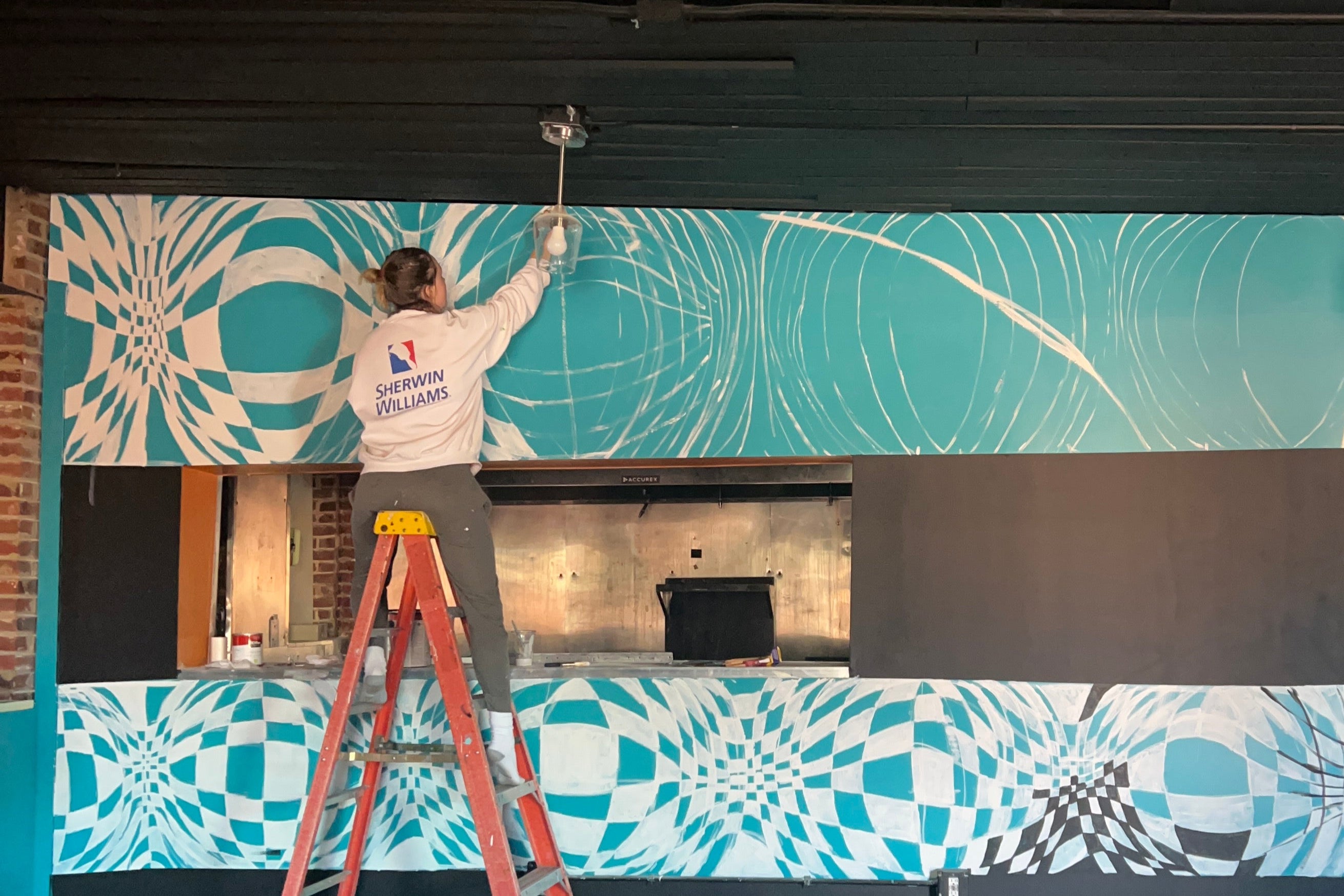 Summer is reaching high on the ladder painting a mural they completed at a restaurant called La Calle in St Louis Missouri. It is halfway painted in this picture. Covering up the teal color behind it. Making a wavy black and white checkered pattern. 
